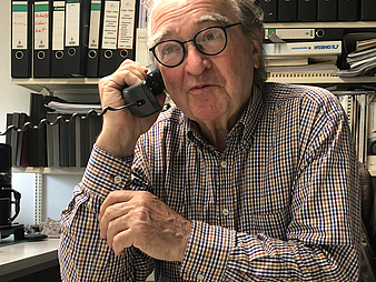 Ein älterer Herr sitzt an einem Schreibtisch und hält sich ein Telefon mit Schnur ans Ohr.