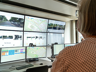Eine Frau vor neun Bildschirmen. Auf ihnen sind unter anderem Landkarten und Fotos von Autos auf der Straße zu sehen.