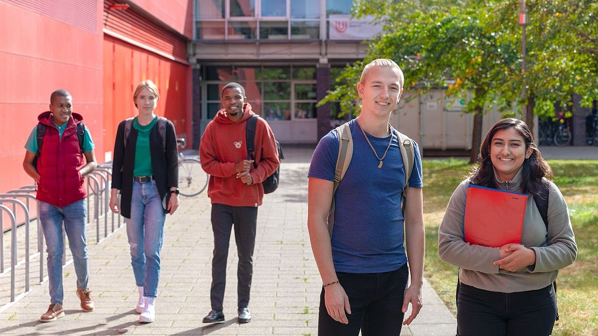 Studierende gehen am Gebäude MZH entlang. Ein junger Mann und eine junge Frau im Vordergrund lächeln in die Kamera