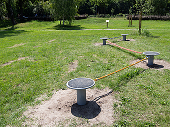 Ein niederschwelliges Balancier-Element auf einer Wiese in einem Park. Mehrere runde Trittplattformen aus Metall sind in kurzer Entfernung voneinander aufgestellt und mit flexiblen, farbigen Balancierstangen verbunden. Die Anlage liegt im Freien und ist von Gras umgeben.