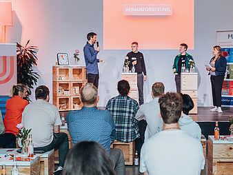 Foto von der StartUp-Lounge: Vier Personen stehen auf einer Bühne, davor sitzt Publikum