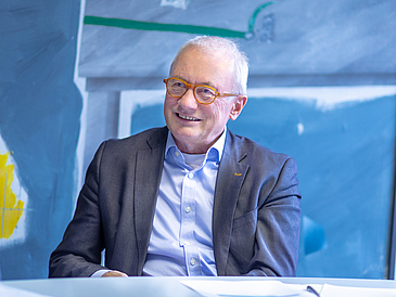 Mann mit Brille sitzt am Tisch vor einem Gemälde und lächelt.