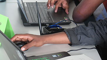 Two students are working on one IoT device. One can see the device, the hands and their computers.