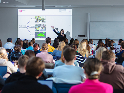 Dozentin vor einer Leinwand und viele Studierende, die zuhören