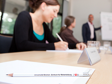 Ein Bleistift mit der Aufschrift "Universität Bremen Zentrum für Weiterbildung" liegt auf einem Tisch in einem vollen Seminarraum.