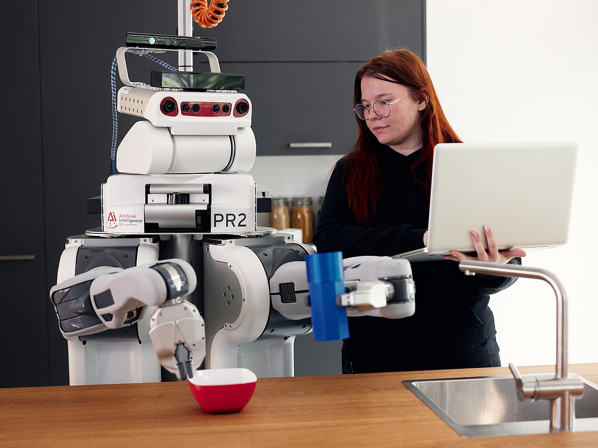 In einer Küche hantiert ein Roboter mit Küchengeräten, neben ihm steht eine Frau mit einem Laptop.