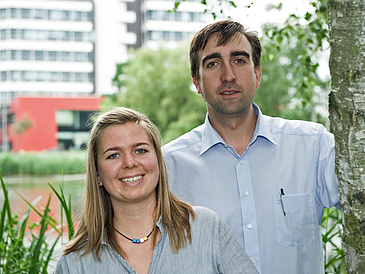 Ein Mann und eine Frau lächeln in die Kamera, im Hintergrund ein Gebäude.