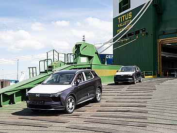 Ein Autoschiff auf dessen Laderampe einige Autos stehen. Diese werden aus dem Autoschiff herausgefahren. Die Autos sind neue Autos, was durch die Schutzfolie auf der Motorhaube sichtbar ist.