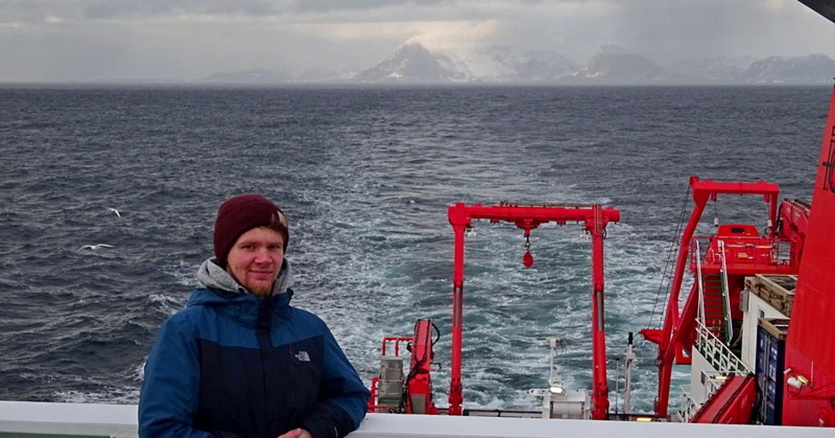A scientist stands on the deck of the research vessel Maria S. Merian wearing a blue and black jacket and wool cap. The stern of the ship and the southern tip of Greenland can be seen in the background.