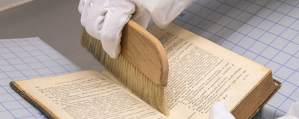 KennenSieSchon_Restaurierwerkstatt_Clebowski Mit einer Bürste geht eine behandschuhte Hand durch die Falz des Buches.