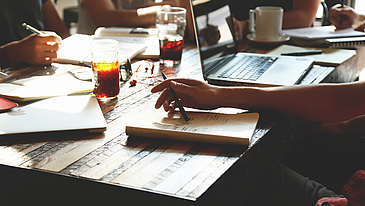 Das Foto zeigt ausschnittsweise einen Holztisch, an welchem Menschen an Laptops und mit Notizbüchern sitzen und arbeiten und Tee trinken. 