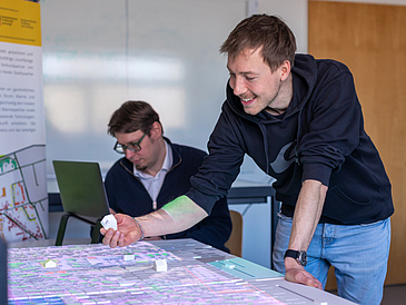 Zwei Personen stehen und sitzen an einem beleuchteten Planungstisch, eine Person hält ein Miniaturhaus in der Hand