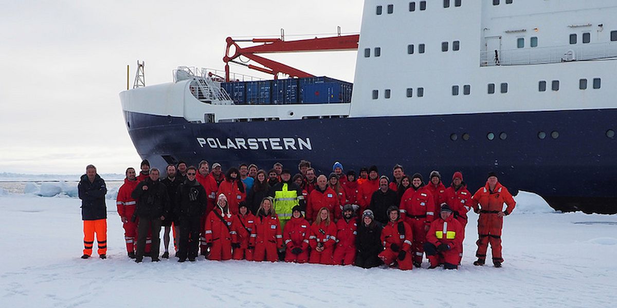 The cruise participants stand for a group photo in red full body suits on an ice floe in front of the research vessel Polarstern.