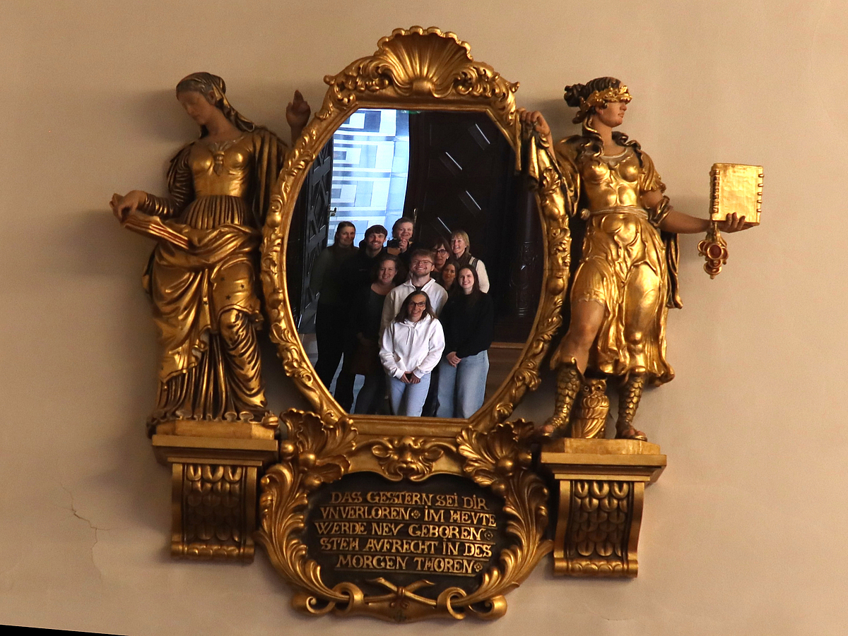 Spiegel-Selfie im Bremer Rathaus Ein goldener Spiegel mit reichen Verzierungen. Links vom Spiegel eine goldene Figur, die in einem Buch liest und die Vergangenheit repräsentiert und rechts vom Spiegel eine goldene Figur mit verbundenen Augen und einem geschlossenen Buch, die die Zukunft repräsentiert. Im Spiegelbild sieht man eine Gruppe, die ein Selfie macht.