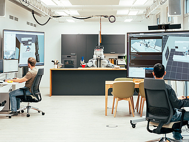 IntEL4CoRo Blick in eine Küche mit Roboter. An den Rändern sind Schreibtische mit Computern zu sehen, an denen Menschen arbeiten.