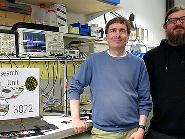 Wissenschaftlicher Leiter Privatdozent Dr. Stefan Bosse (links) der Universität Bremen und Doktorand M.Sc. Physik Christoph Polle (rechts) vom Faserinstitut Bremen stehen im Labor vor der Messanlage und dem Prototypen einer sensorierten Faser-Metall-Laminat Platte.