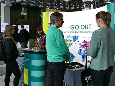 Mann im Gespräch mit einer Studentin vor dem Infostand