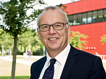 Professor Bernd Scholz-Reiter steht im Campuspark und im Hintergrund sieht man das MZH-Gebäude.