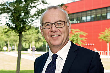 Professor Bernd Scholz-Reiter steht im Campuspark und im Hintergrund sieht man das MZH-Gebäude.