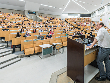 Ein Lehrender spricht vor Zuhörern im vollen Hörsaal.