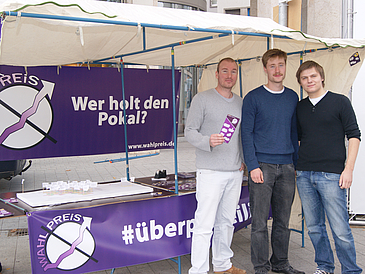 Drei Männer vor einem Infostand.