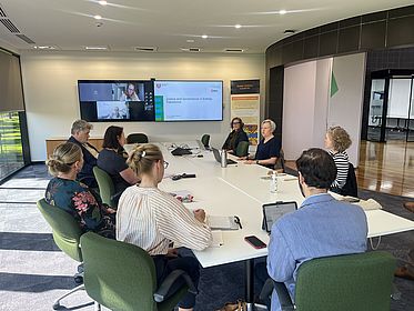 In einem Konferenzraum sitzen Menschen um einen Tisch herum und hören Stefanie Baaschs Vortrag, auf Bildschirmen an der Wand sind weitere online-Teilnehmende zu sehen.