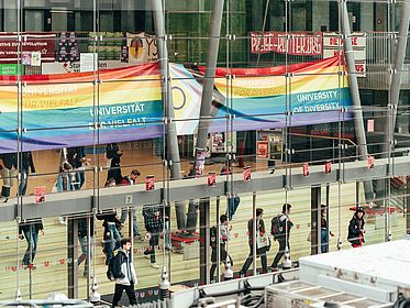 Menschen in der Glashalle, Pride-Flagge im Hintergrund.