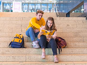 Young man and woman are sitting on stairs and are looking together in a book.