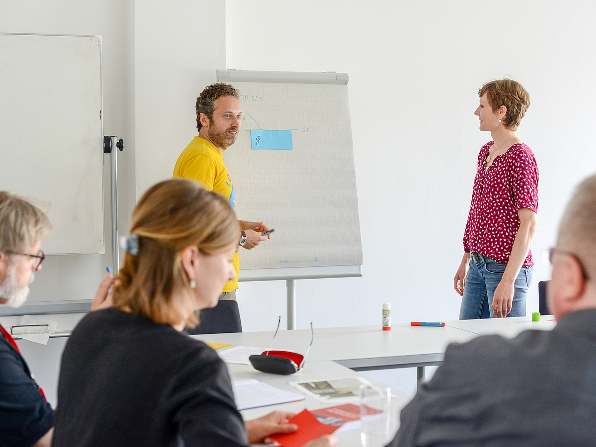 Zwei Lehrende arbeiten vor Zuhörern an einem Flipchart.