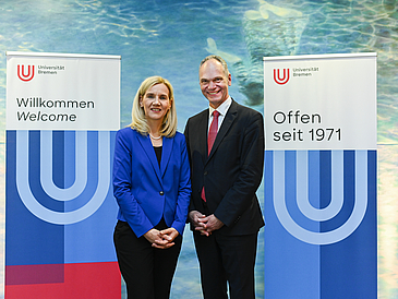 Freuen sich über die NominieRektorin der Universität Bremen, Professorin Jutta Günther, und Präsident der Carl von Ossietzky Universität Oldenburg, Professor Ralph Bruder, stehen zwischen Aufstellern der Uni Bremen und lächeln in die Kamera.