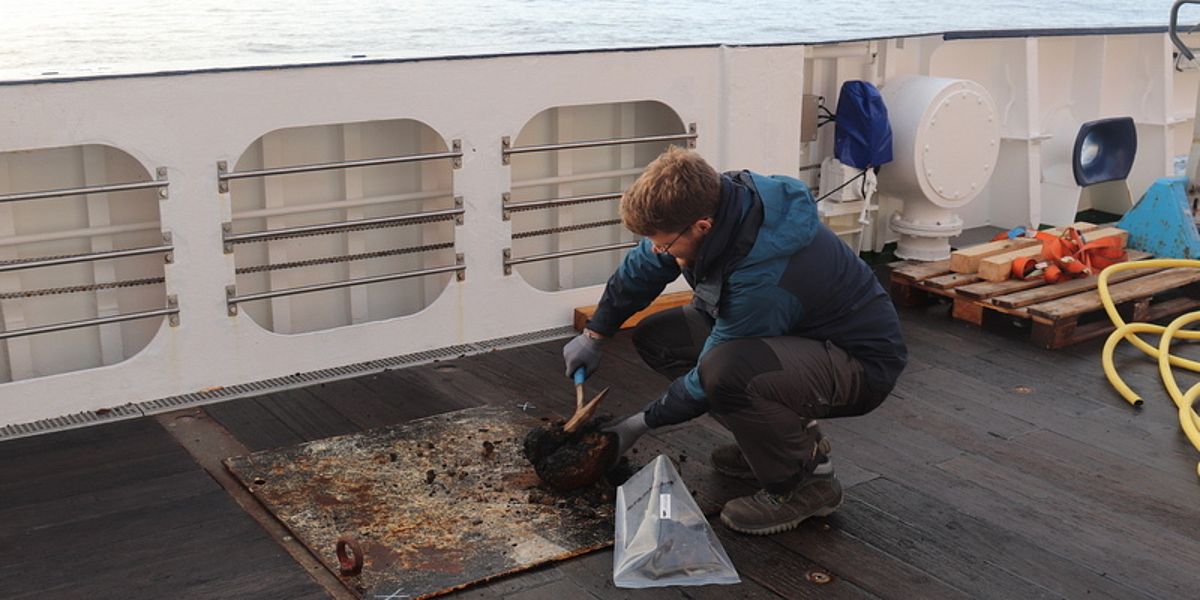 Ein Wissenschaftler hockt nahe der Reeling an Deck des Forschungsschiffes Maria S. Merian und zerkleinert Gesteinsproben mit Hilfe eines Geologenhammers.