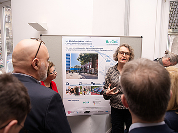 Foto der Poster-Session, bei der eine interessierte Besucherin Fragen zu dem Arbeitspaketen stellt. Auf diesem Foto handelt es sich um das Poster der Hochschule Bremen zu dem Thema “Mit Modellprojekten zu einer Campusentwicklungsstrategie“ das von der Leiterin des Projektes vorgestellt und erklärt wird.