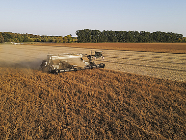 Eine Landwirtschaftsmaschine im Einsatz auf einem Feld. Im Hintergrund sieht man Wald und blauen Himmel.