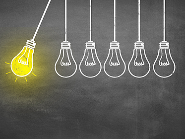 AdobeStock_jd8Fih.jpg Vor einer schwarzen Tafel sieht man wie mit Kreide gezeichnete Glühbirnen, die auf derselben Höhe herunter hängen. Sie sind wie ein Kugelstoßpendel angeordnet: links holt eine gelb leuchtende Glühbirne aus, die anderen fünf hängen ohne Leuchteffekt rechts daneben.