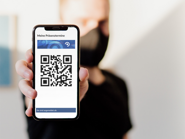 Junger Mann mit Alltagsmaske zeigt Handy mit QR-Code