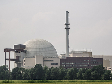 Ansicht des Reaktorgebäudes und Schornstein des Atomkraftwerks Brokdorfs vor grauem Himmel, im Vordergrund eine grüne Wiese und Laubbäume