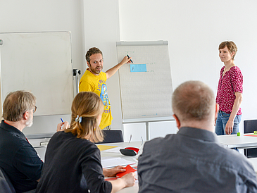 Zwei Lehrende arbeiten vor Zuhörern an einem Flipchart.