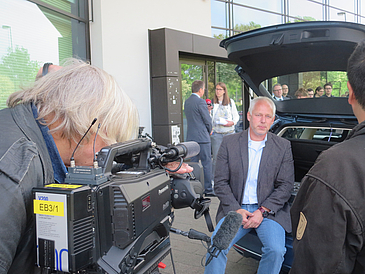 Mann sitzt am offenen Kofferraum und wird von Fernsehteam interviewt