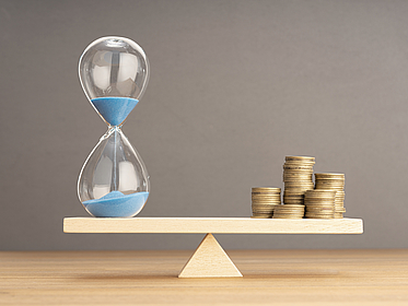 Balance zwischen Zeit und Geld Konzept. Sanduhr und gestapelte Münzen auf Wippe. Balance zwischen Zeit und Geld: Verdeutlicht durch Sanduhr und gestapelte Münzen auf Wippe