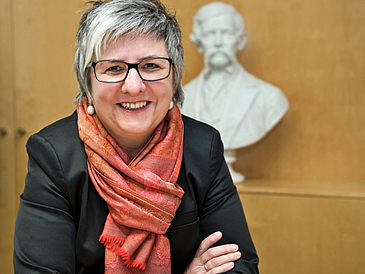 Portrait einer Frau mit kurzen grauen Haaren und Brille. Sie trägt ein schwarzes Jacket mit einem roten Schaal.