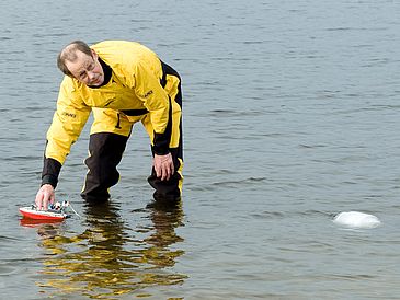 Mann in Trockenanzug steht knietief im Wasser während ein Spielzeugboot versucht, einen Eisblock zu ziehen.