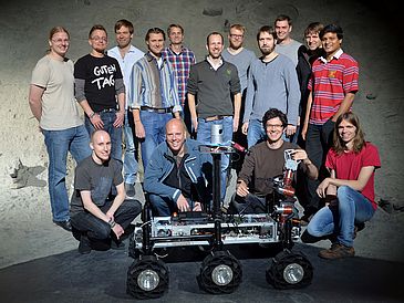 Gruppenbild mit 15 Männern und einem Roboter vor einer künstlichen Mondlandschaft.