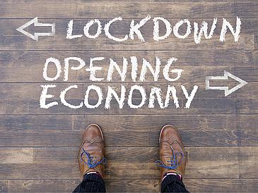 Eine Person steht vor einem Kreideschriftzug auf dem Boden: "Lockdown" mit einem Pfeil nach links, "Opening Economy" mit einem Pfeil nach rechts