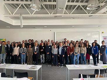 Gruppenfoto: Etwa 40 Personen stehen in zwei Reihen hinterainder in einem Seminarraum