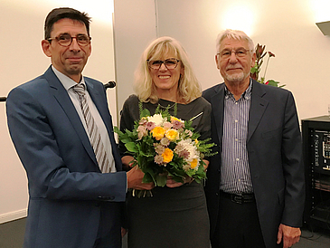 zwei Männer und eine Frau mit Blumenstrauß stehen im Halbkreis