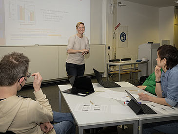 Im Hintergrund eine vortragende Frau vor einer Präsentation, im Vordergrund ein Gruppenarbeitstisch