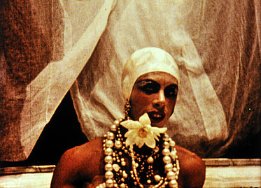 Person mit Kopfhaube und viel Schmuck vor Hintergrund mit Gardinen