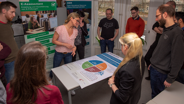 Eine Gruppe aus Lehrenden und ZMML-Projektteam stehen um einen Tisch und schauen sich ein Poster an