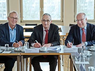 Drei Männer sitzen am Tisch und unterzeichnen ein Dokument