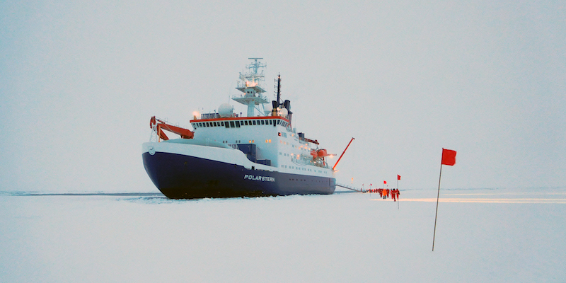 2016: PS101 - ARK XXX-3 (Karasik-Seamount) Das Forschungsschiff Polarstern liegt an einer Eisscholle vertäut, die Gangway ist ausgefahren, Fahrtteilnehmer stehen im Hintergrund in roten Ganzkörperanzügen auf dem Eis, im Vordergrund sind Stangen mit roten Fahnen zu erkennen, die alle paar Meter ins Eis gesteckt wurden.